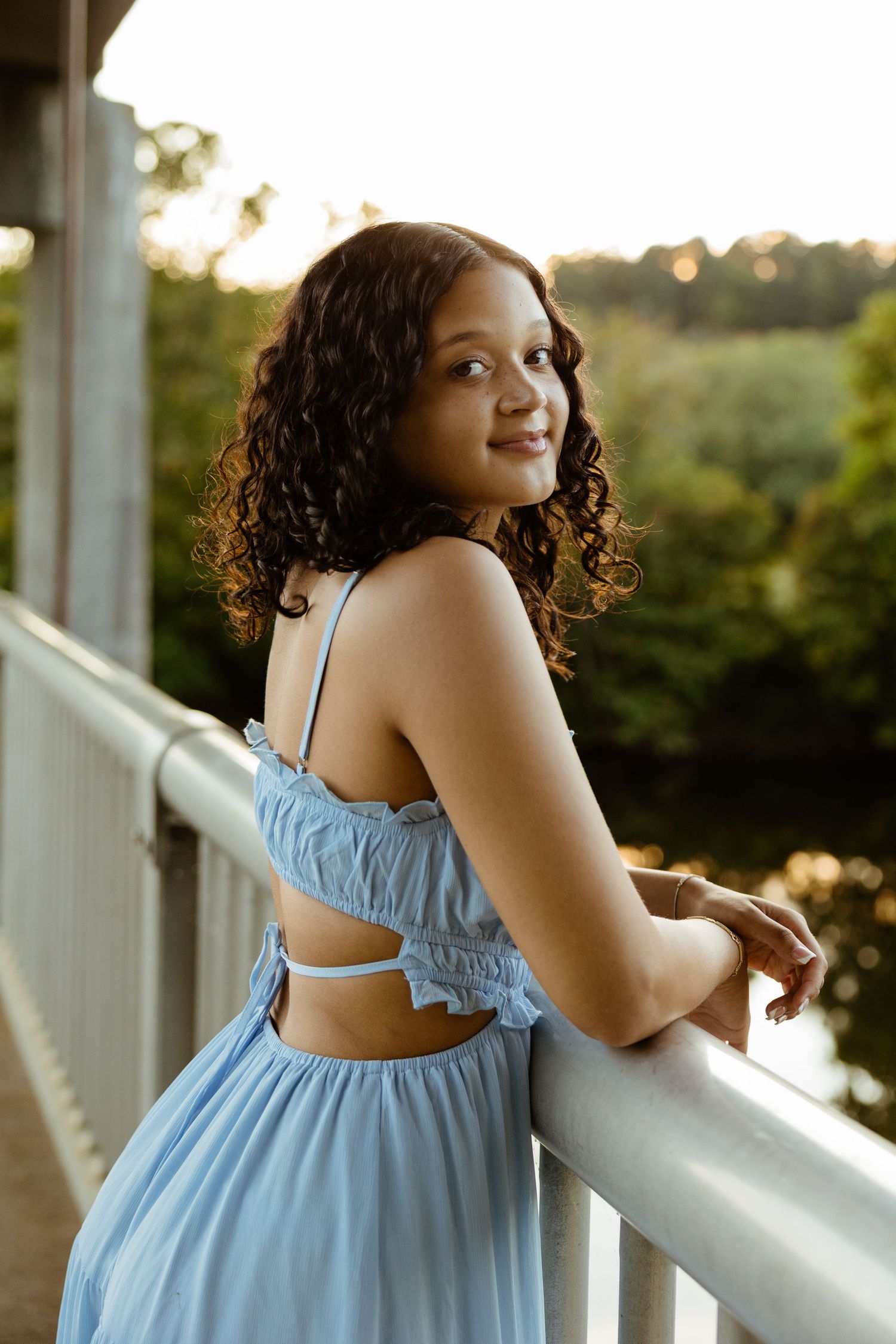 A smiling young woman in a light blue dress leans on a railing during a golden sunset portrait session.