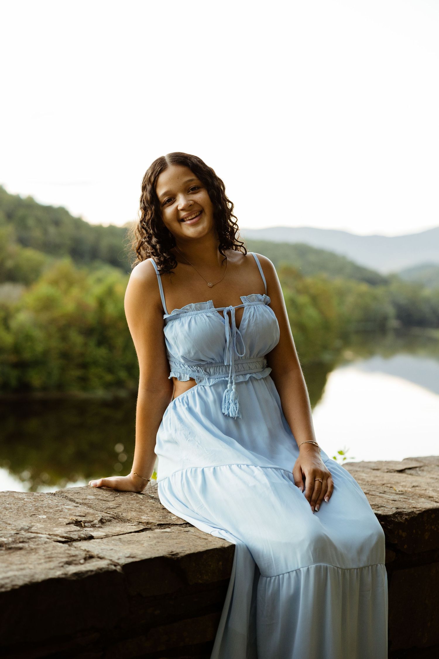A cheerful young woman in a blue two-piece dress sits on a stone wall with a scenic river backdrop.