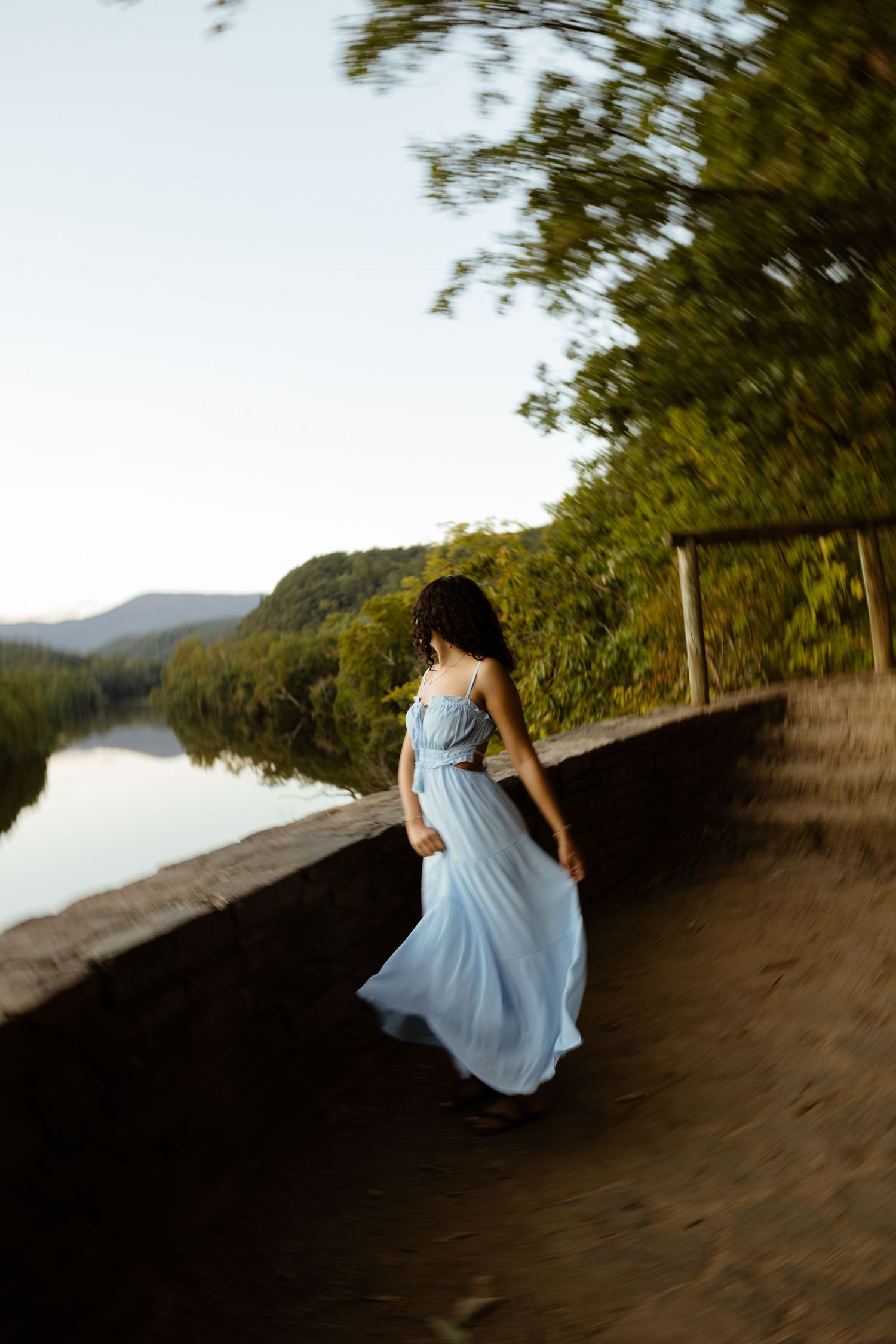 A young woman in a flowing light blue dress walks along a scenic riverside path at golden hour.