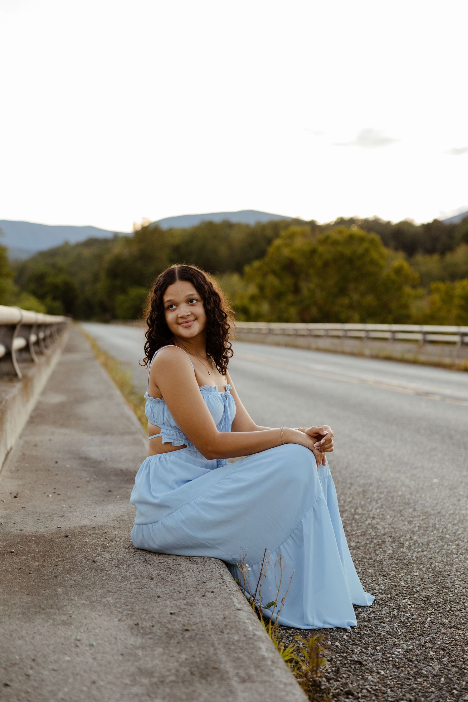 A smiling young woman in a light blue dress sits on a bridge ledge with mountain scenery behind her.