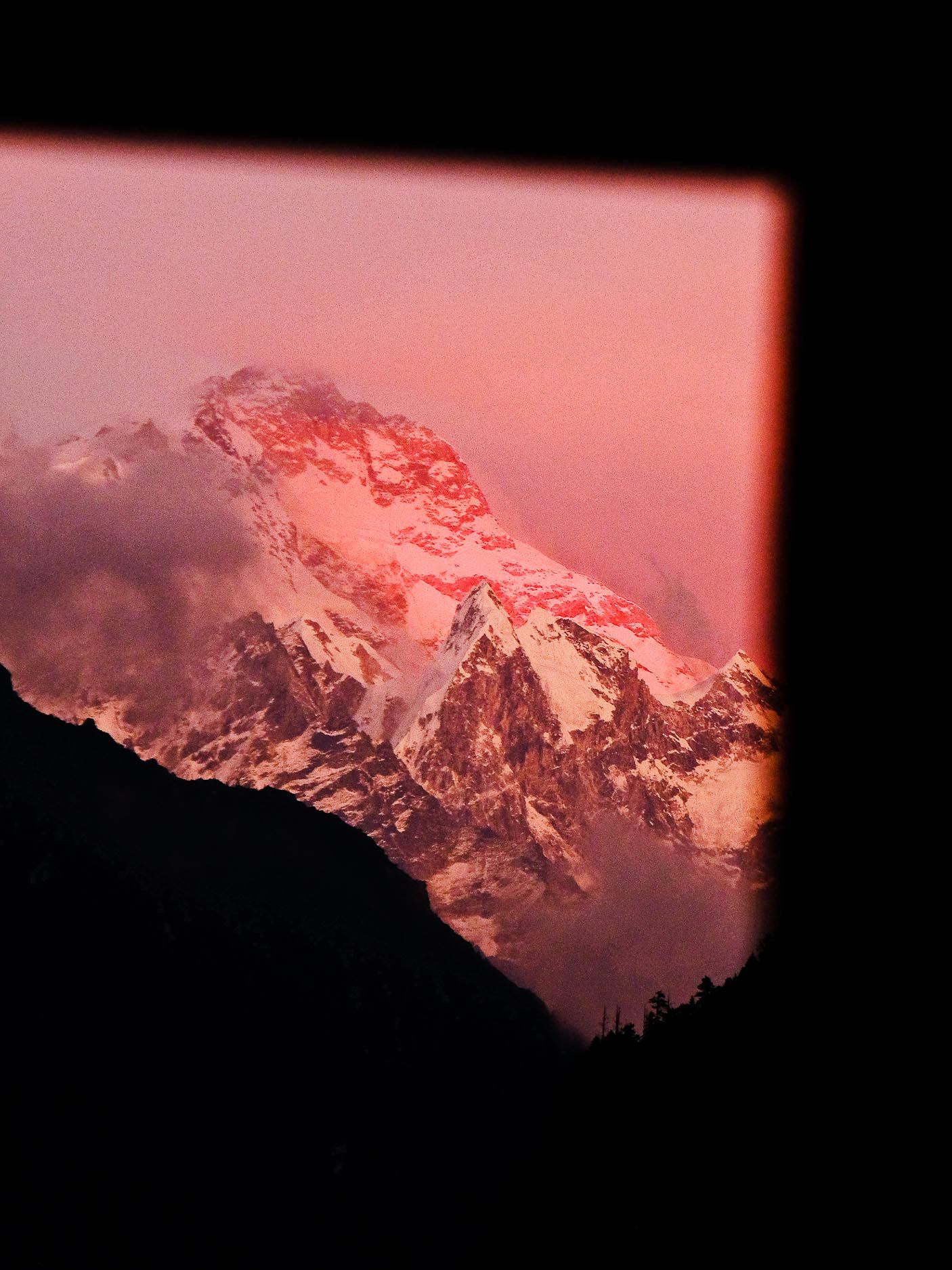 Snow-covered mountain peak bathed in pink alpenglow viewed through a window at sunset.