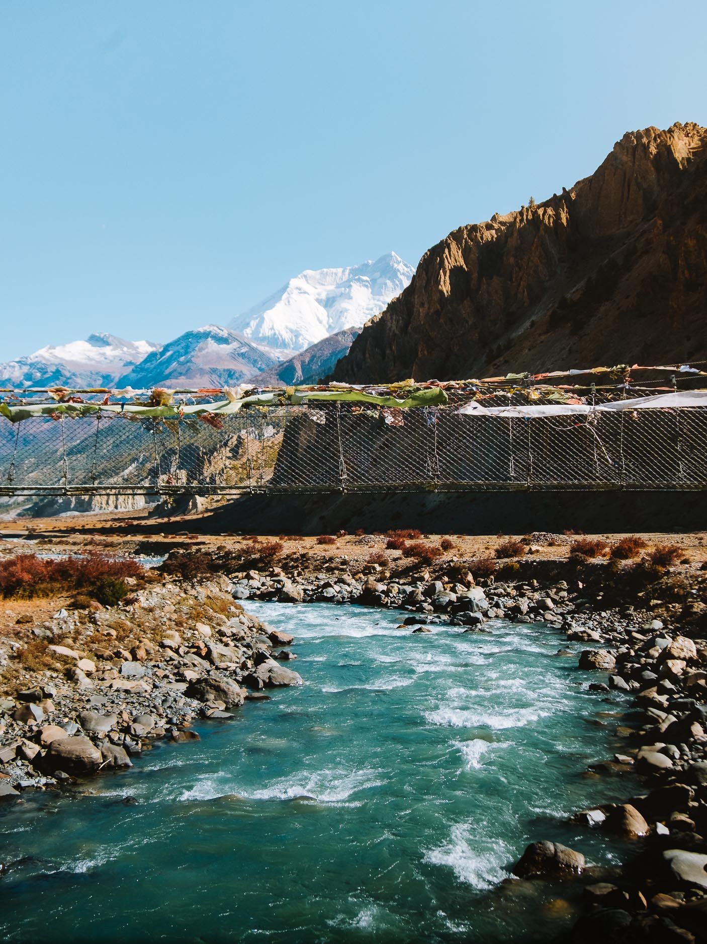Suspension bridge spans a turquoise river with snow-capped mountains in the background.