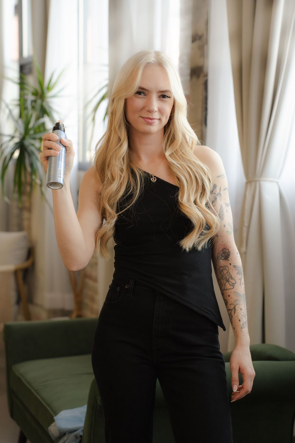 Woman in black outfit holding a spray bottle stands in front of curtains and green couch in a bright interior setting.