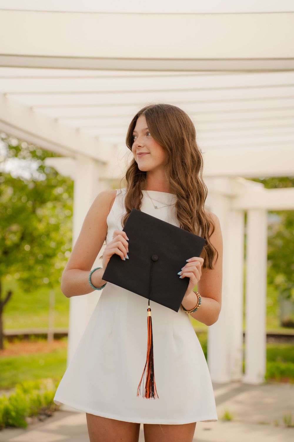 Young woman in white dress holding black graduation cap with tassel, smiling outdoors under white pergola.
