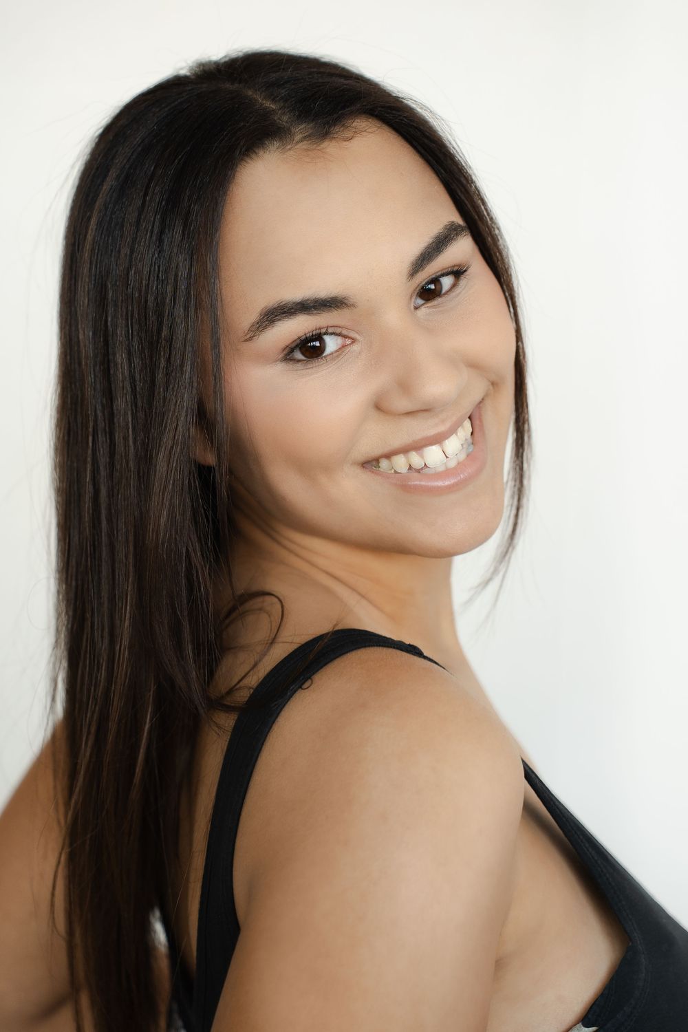Portrait of a young woman with long dark hair wearing a black tank top smiling warmly against a light background.