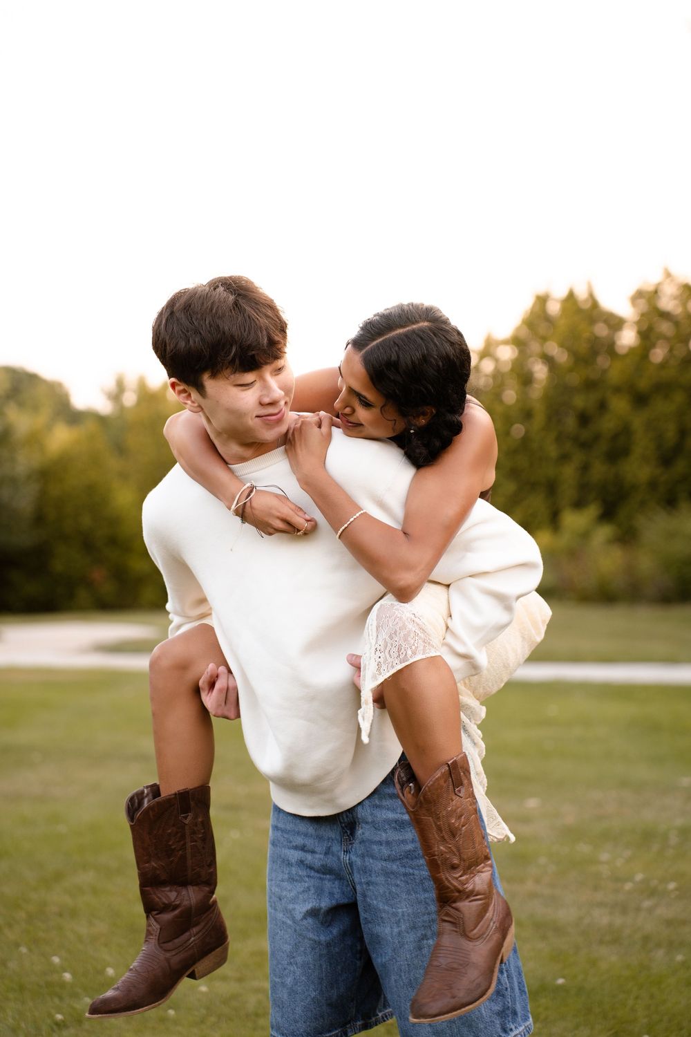 Playful couple in casual attire and cowboy boots share a piggyback ride in a grassy field.