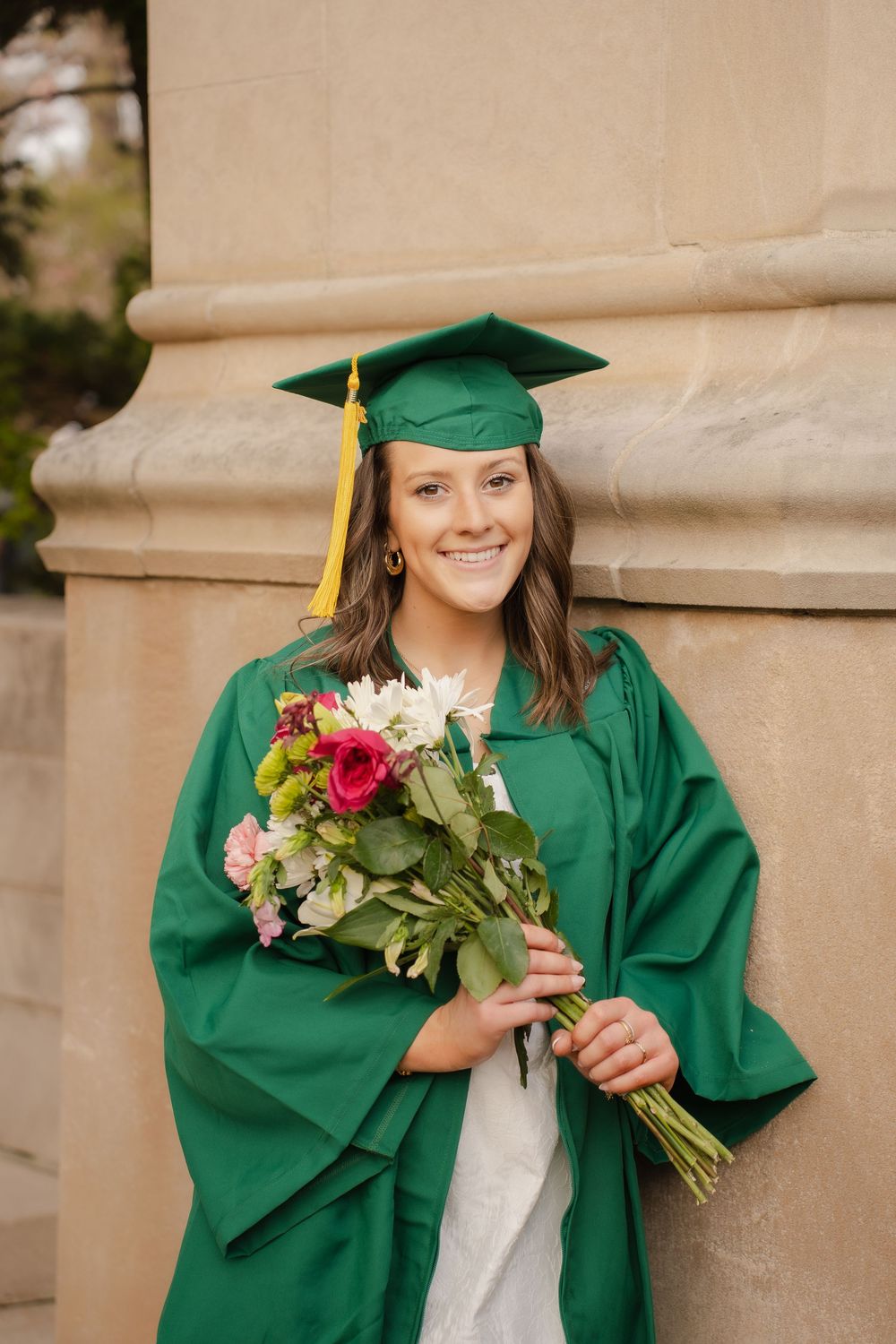 Smiling graduate in green cap and gown holding colorful flower bouquet, posing outdoors near stone column.
