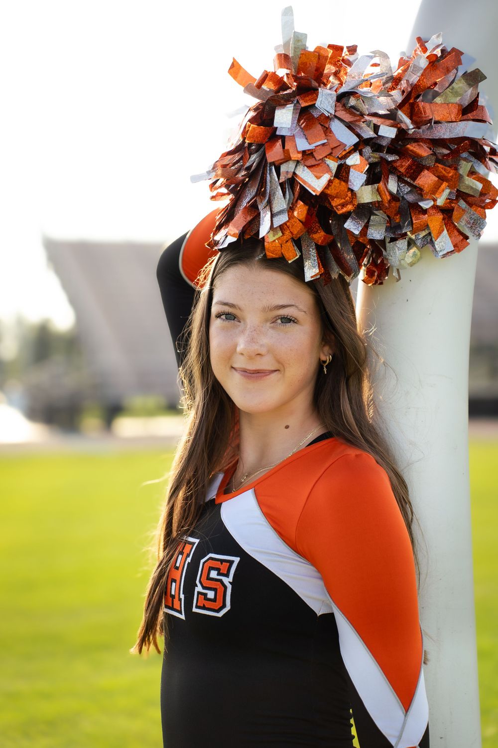 Cheerleader in orange and black uniform with pom-pom hair accessory poses outdoors.