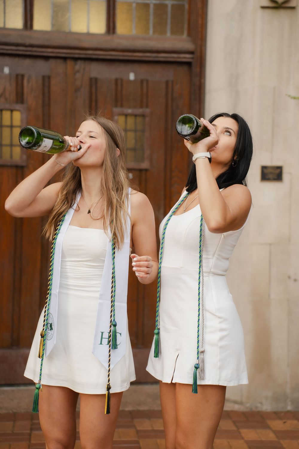 Two young women in white dresses with honor cords drinking from green bottles outdoors near a building entrance.