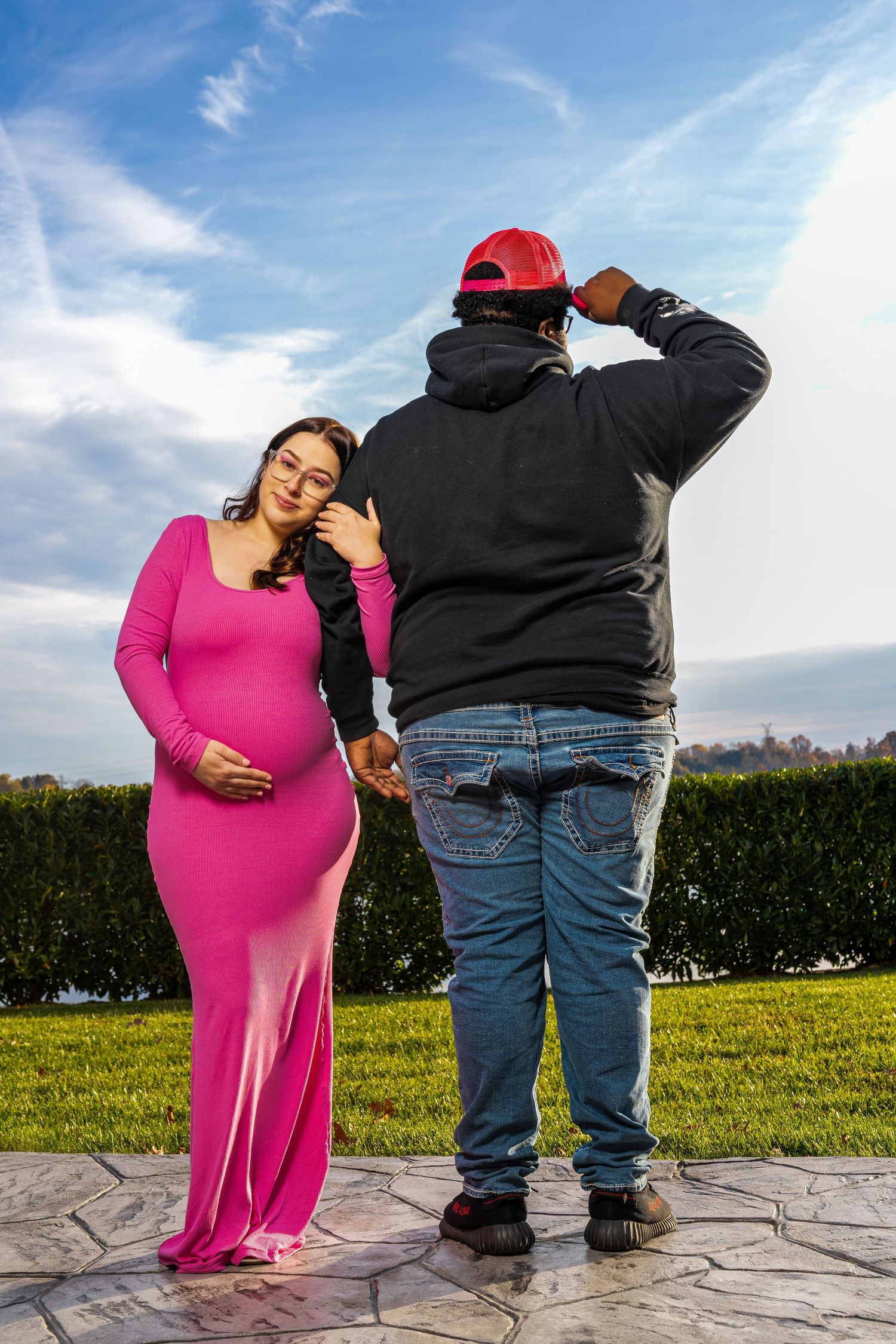 A photographer takes maternity photos of someone in a bright pink dress outdoors.