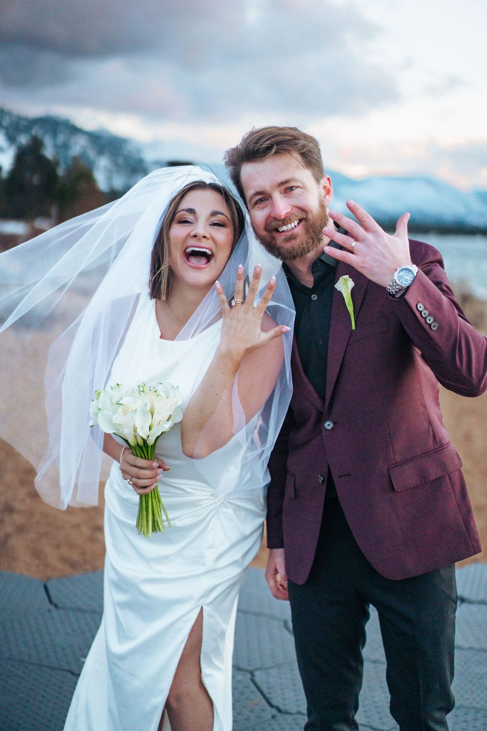 Newlyweds celebrate joyfully showing off rings in burgundy suit and white dress outdoors.