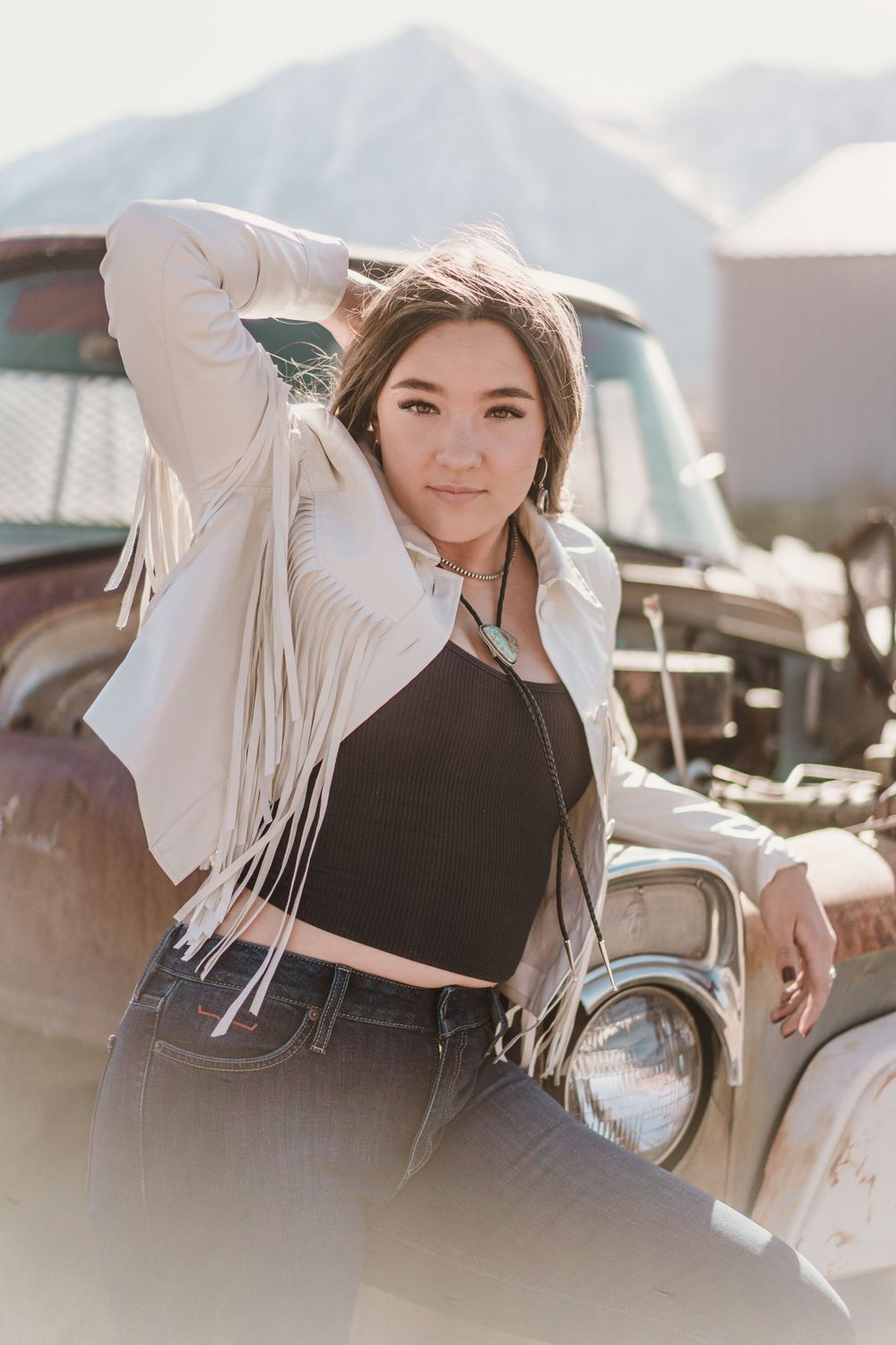 A stylish portrait against a vintage car and mountain backdrop featuring a white fringed jacket and black crop top.