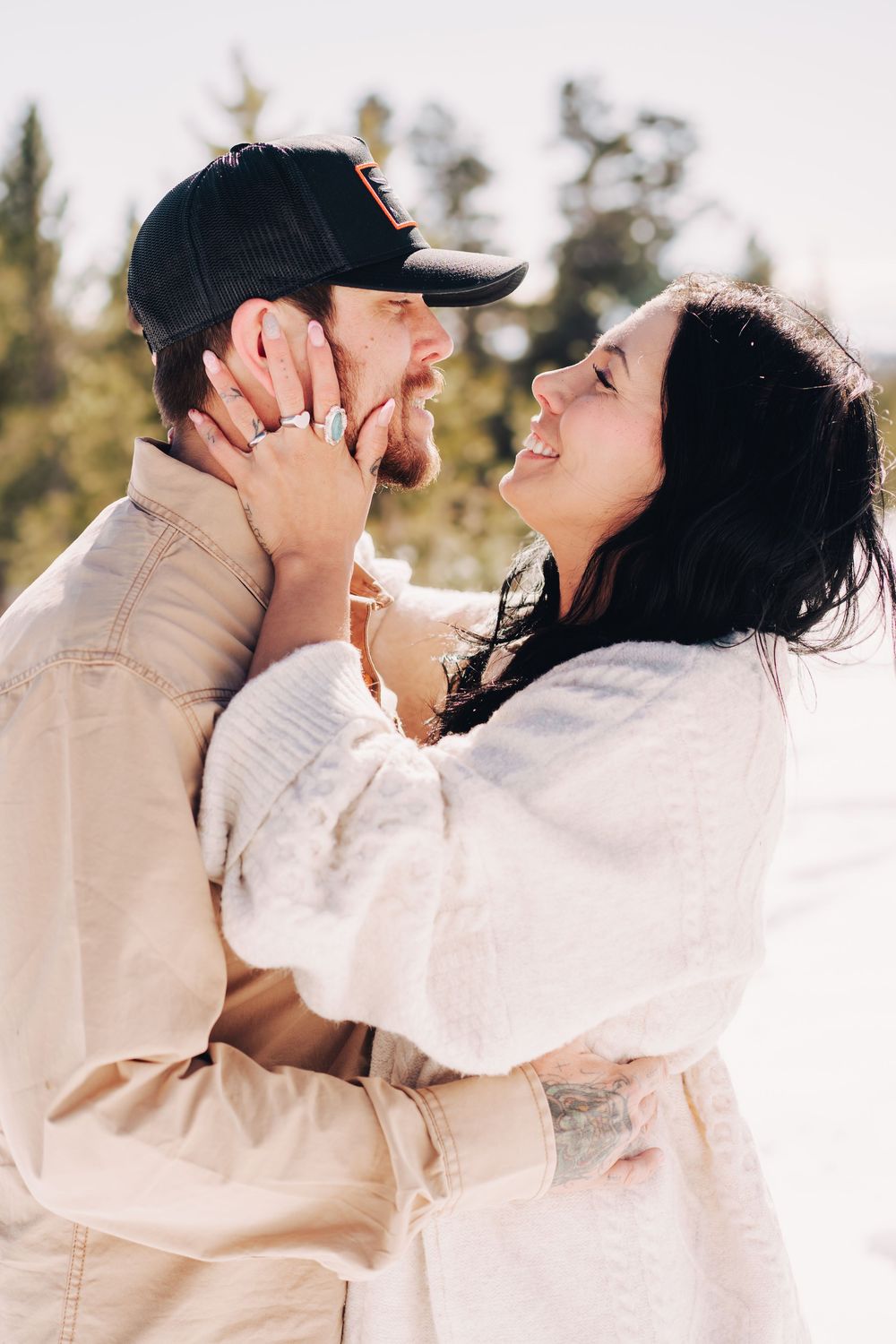 A couple shares a romantic embrace outdoors, woman in white wrap smiling joyfully at bearded man in black cap.
