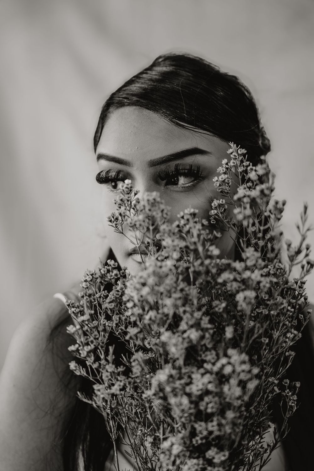 Black and white portrait of a woman holding wildflowers covering her face, with expressive eyes visible above the blooms.