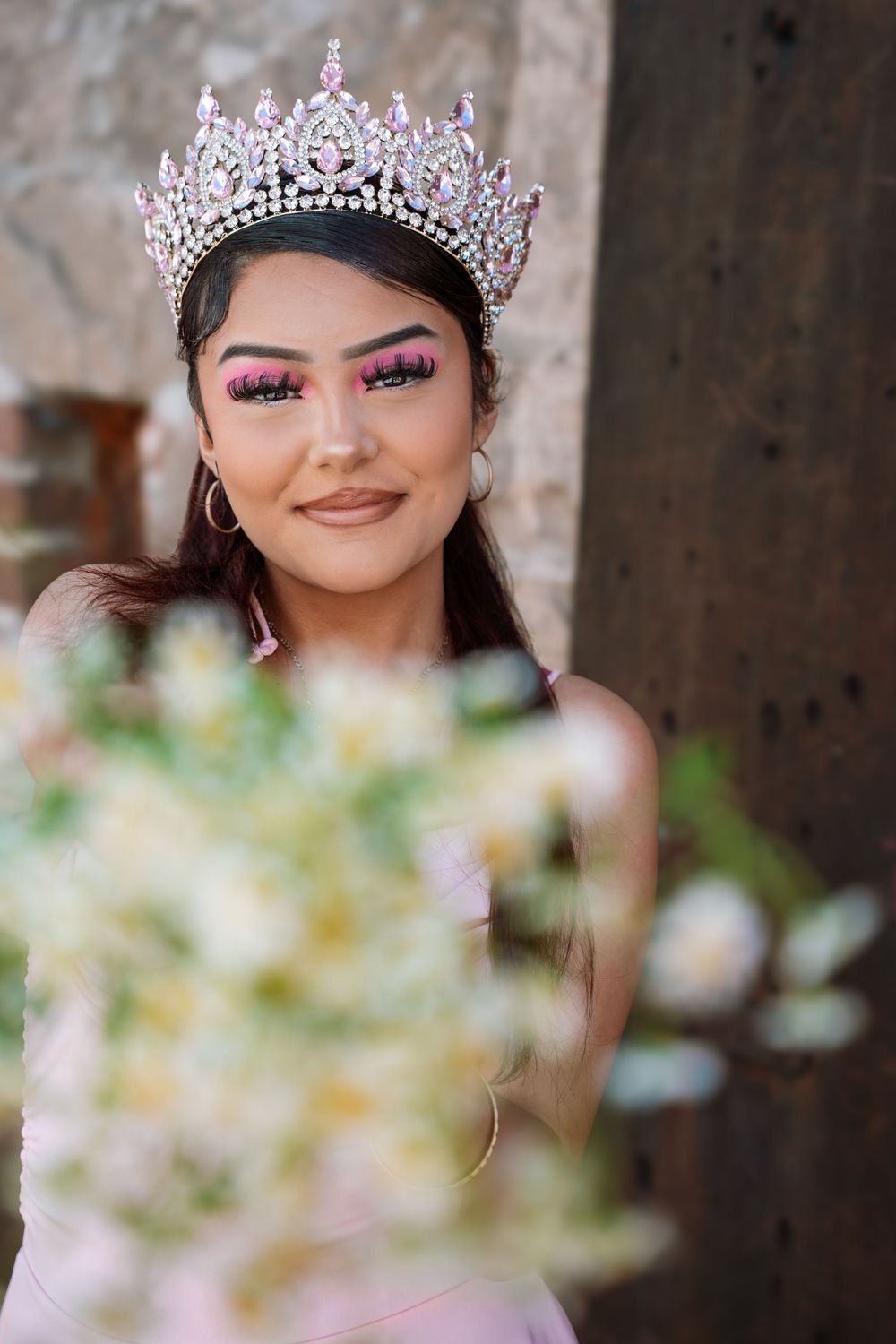 Young woman wearing a sparkling pink tiara crown and bold pink makeup, holding white flowers, in an elegant portrait.