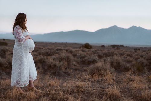 Silhouette of pregnant woman in white lace dress standing in desert landscape at dusk with mountains behind.