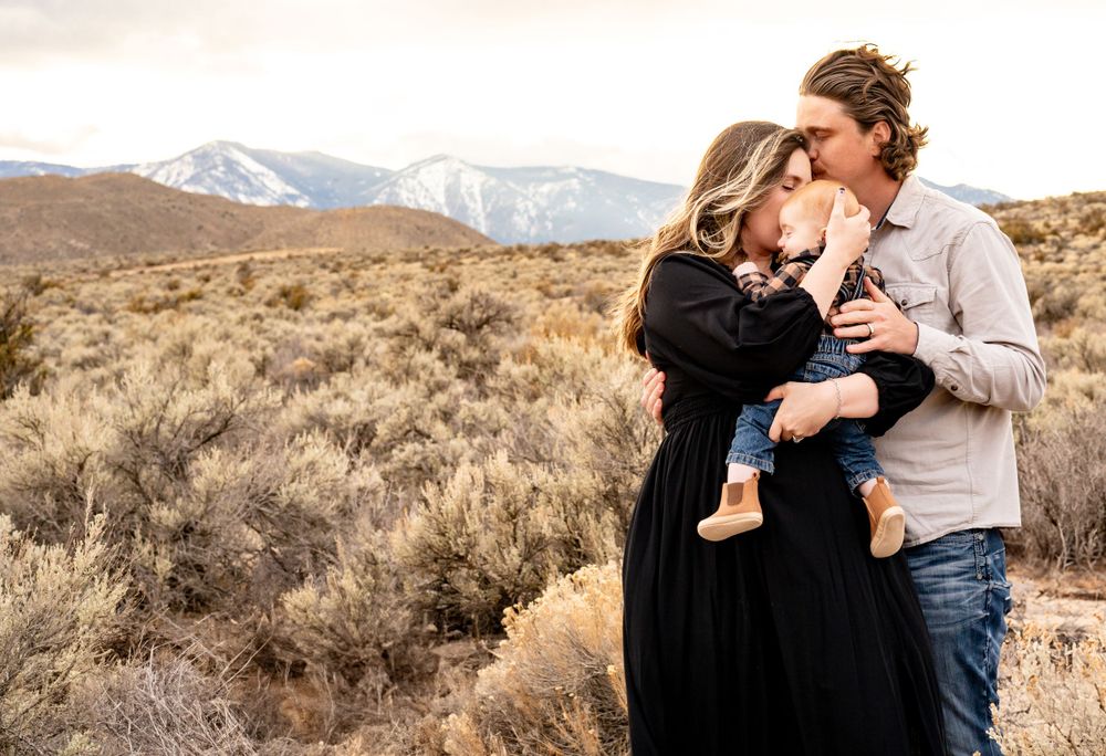 Couple with baby poses against desert mountain landscape during sunset photoshoot.