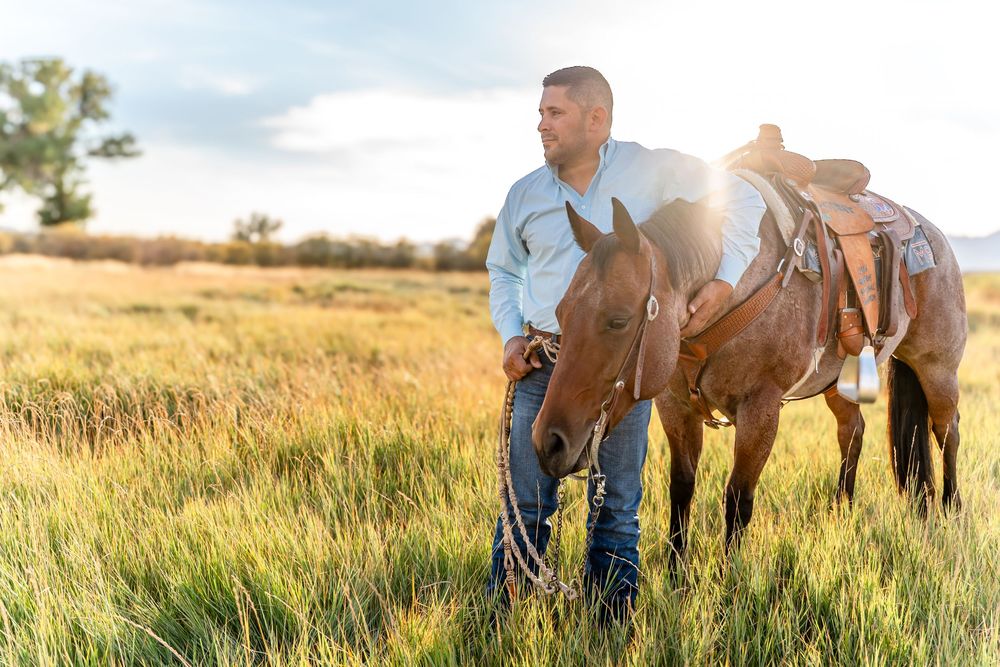Cowboy standing in golden field with saddled brown horse at sunset.