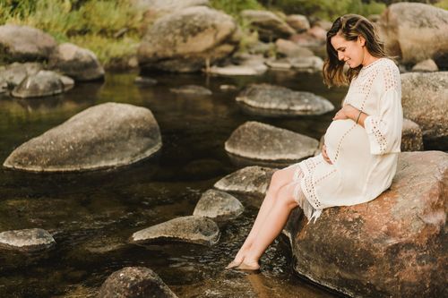 Serene maternity portrait by river with woman in white dress sitting on rocks near flowing water.
