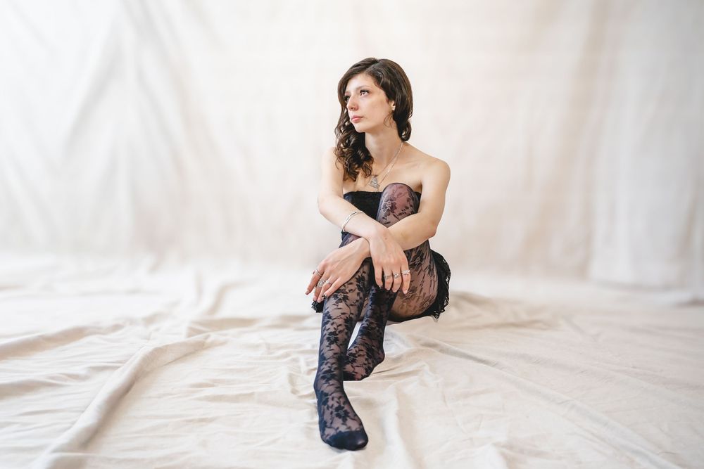 Woman in black lace corset and stockings sitting gracefully on white draped fabric in a studio setting.