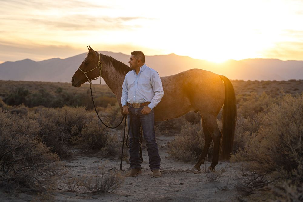 Cowboy standing beside a dark horse at sunset in a desert landscape with mountains in the background.