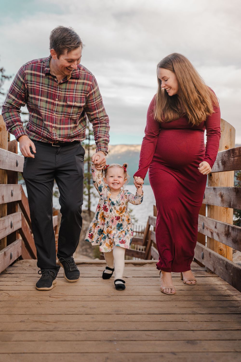 Happy family with toddler girl walking on wooden bridge, pregnant mother in red dress, outdoor maternity photo.
