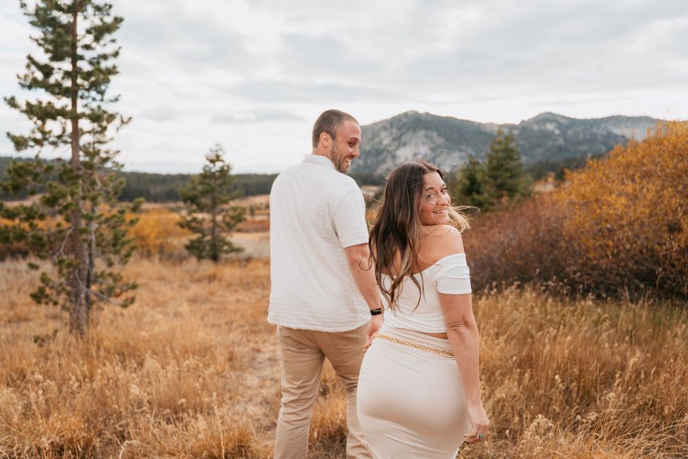 Couple in white outfits holding hands in autumn mountain meadow, woman smiling back at camera.