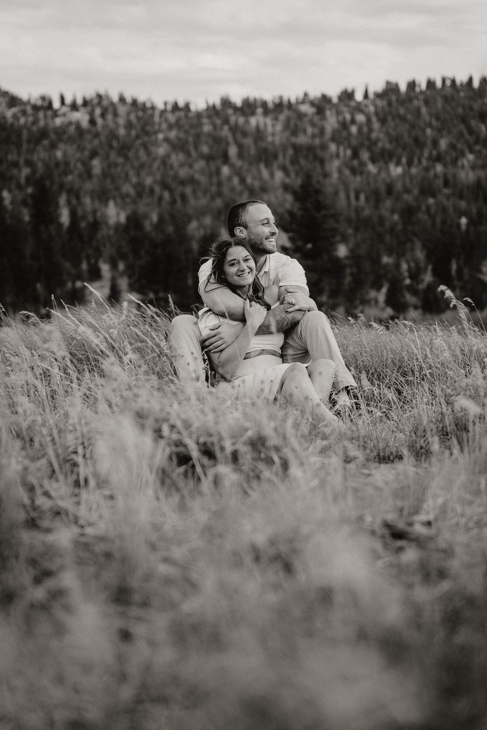 Black and white photo of a couple embracing in a field of tall grass with a forest backdrop.