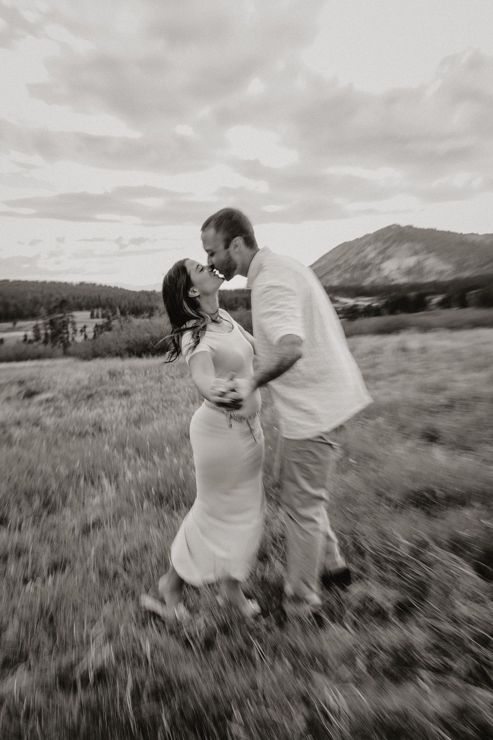 Black and white photo of couple kissing in a meadow, woman in white dress, mountains visible in the scenic background.