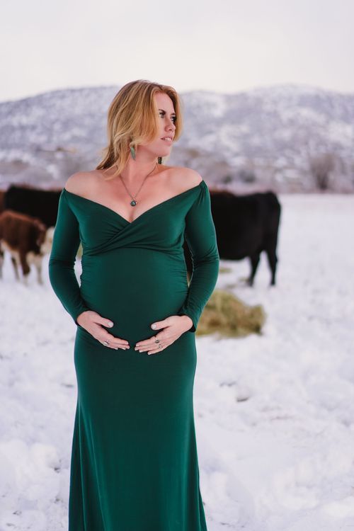 Winter maternity portrait in emerald green dress with snow-covered mountains and cattle in background.