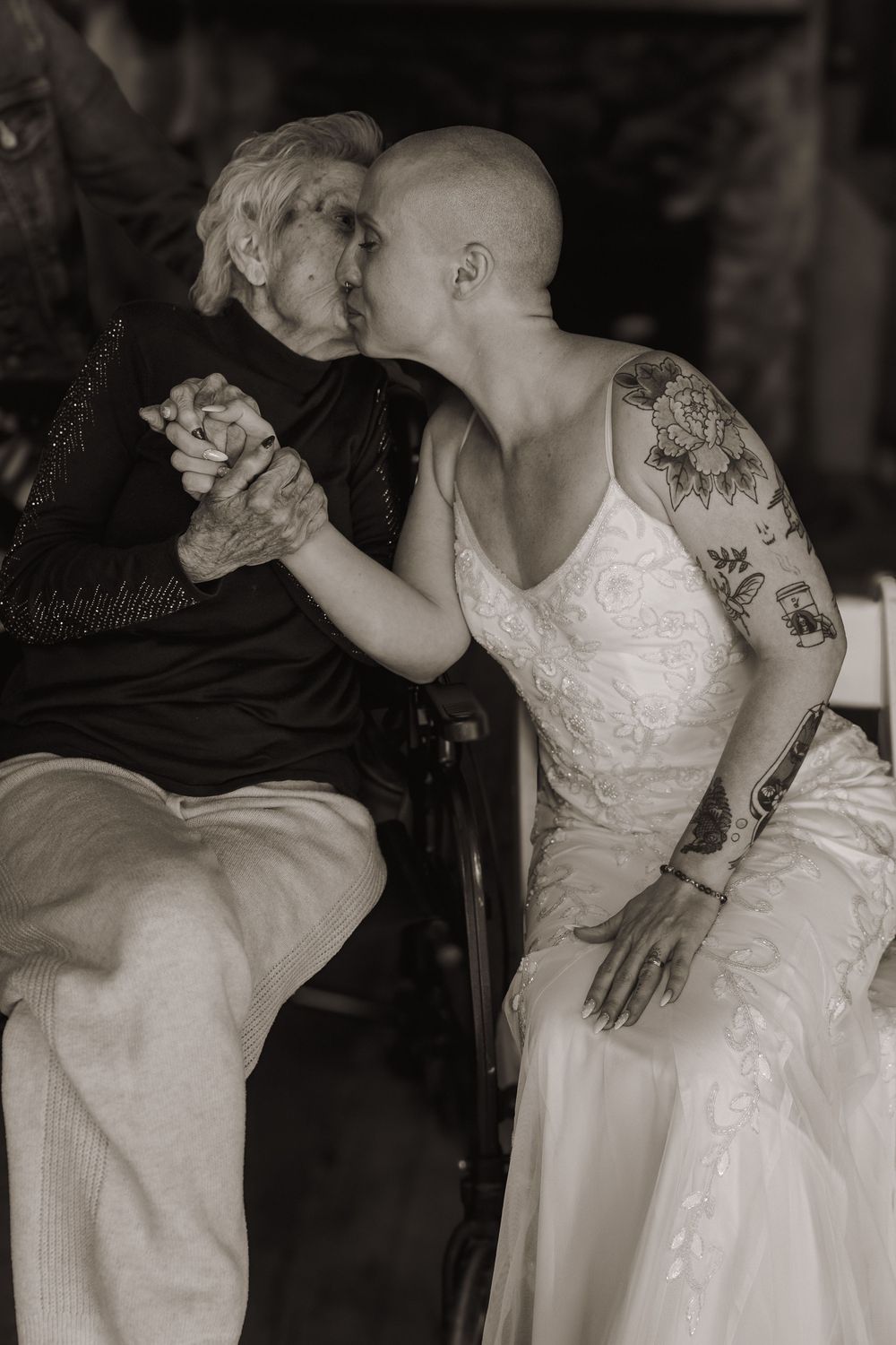 Bald bride in white gown with tattoos kisses elderly woman in black outfit, sepia-toned wedding photo.