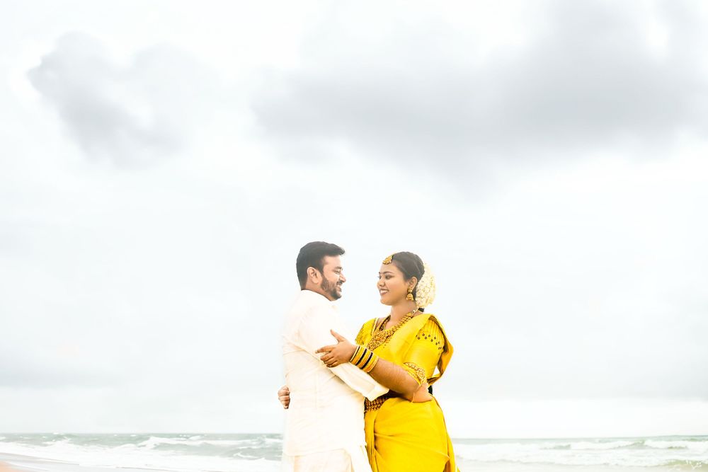 A couple in white and yellow attire embraces on a cloudy beach.