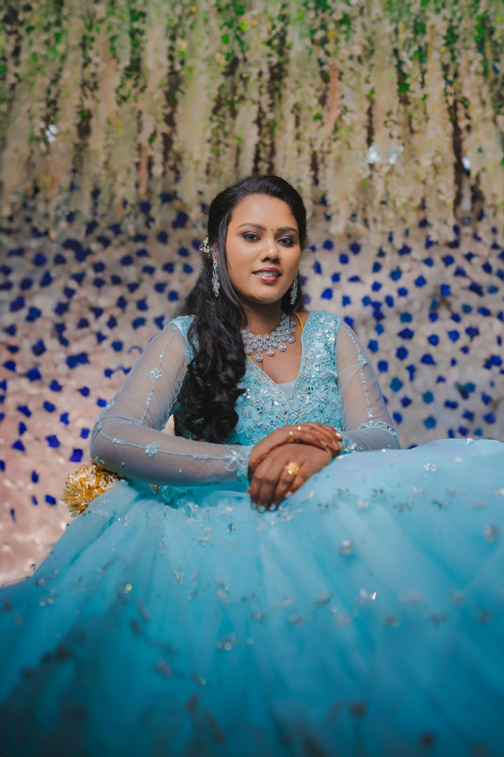 A person in a sparkling blue gown poses against a decorative backdrop with hanging floral elements.