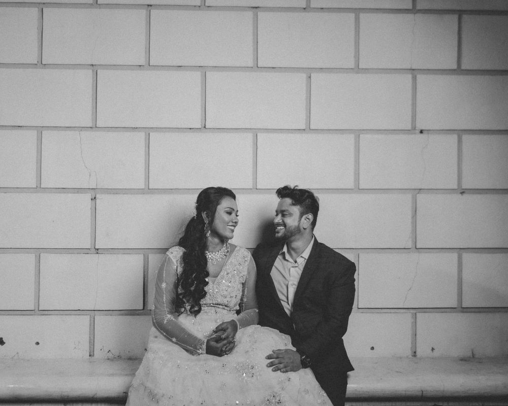 A couple in formal attire sits together on a bench against a white brick wall in black and white.