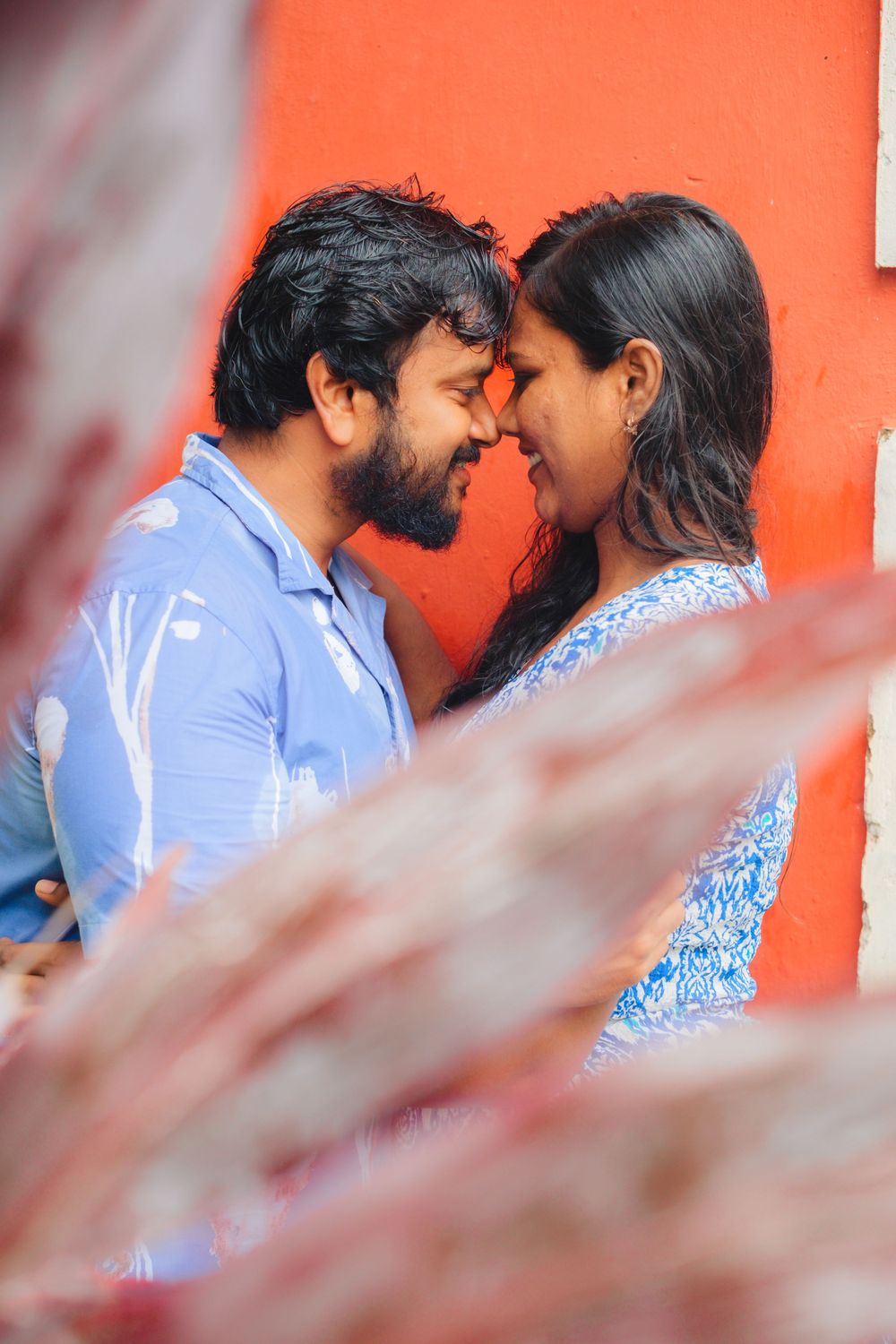 Couple sharing an intimate moment against vibrant orange wall with foliage in foreground.