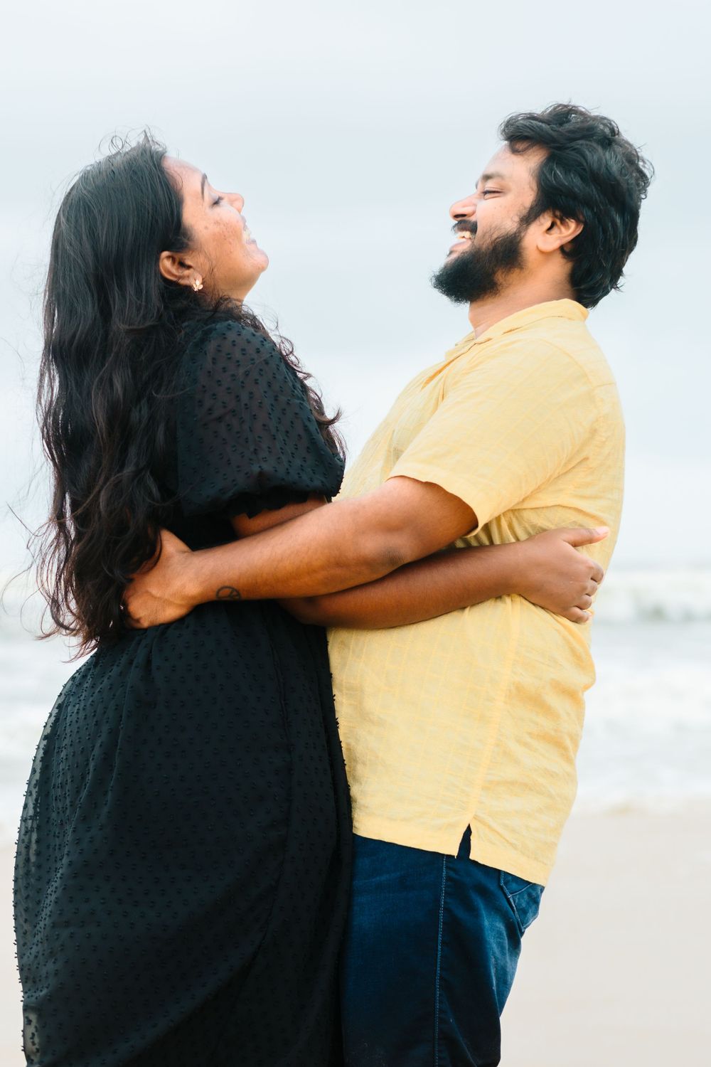 Playful couple embracing and laughing on beach wearing yellow and black outfits.