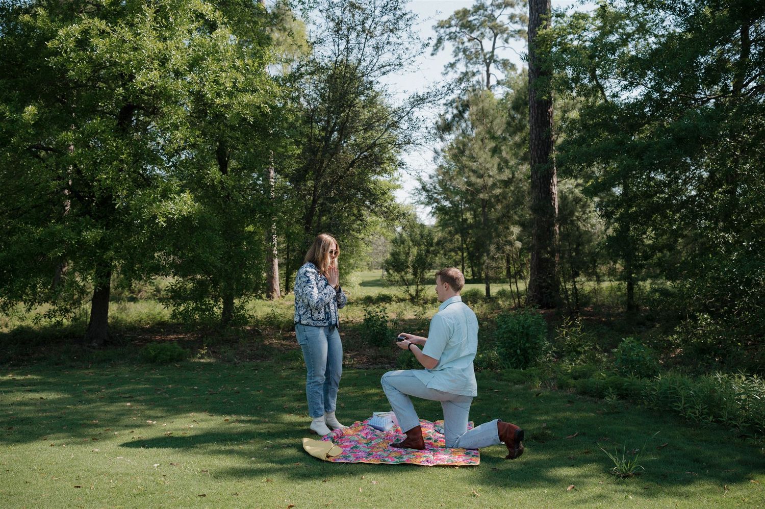 Two people share a romantic moment on a picnic blanket surrounded by lush green trees in a park setting.