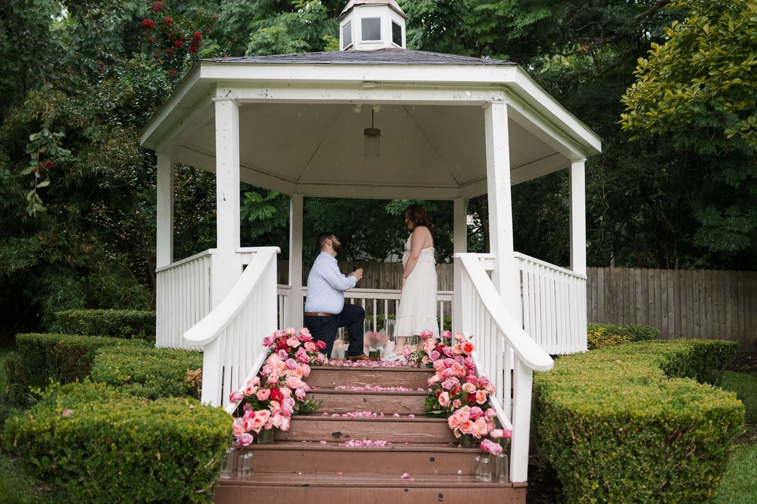 Two people share a romantic moment on a picnic blanket surrounded by lush green trees in a park setting.