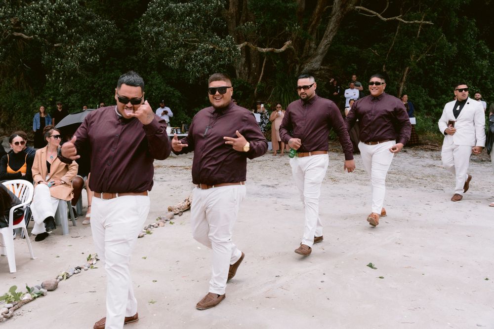 Four groomsmen in matching burgundy shirts and white pants walk in sync on a beach path.