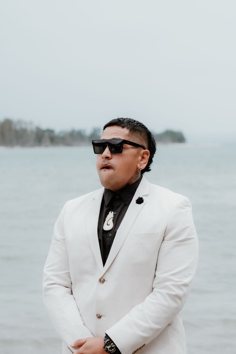 Person in white blazer and black turtleneck poses against ocean backdrop wearing sunglasses.