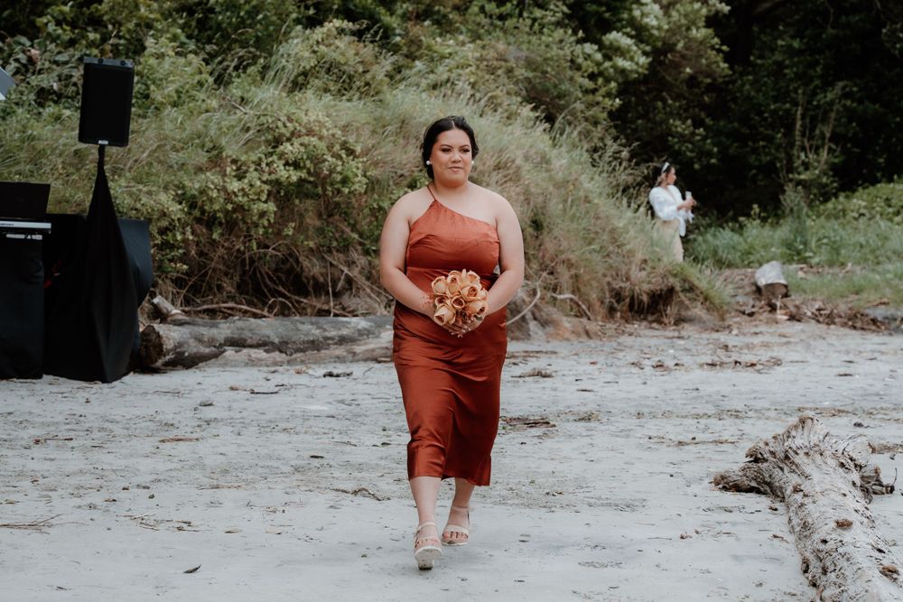Person in rust-colored dress walks along beach holding bouquet.