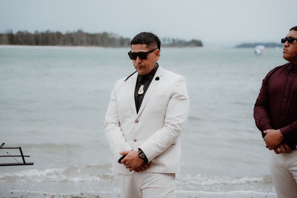 Wedding party member in white suit stands on beach with water and boats in background.