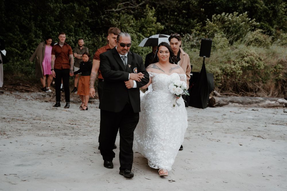Wedding procession walking down sandy path with guests and greenery in background.