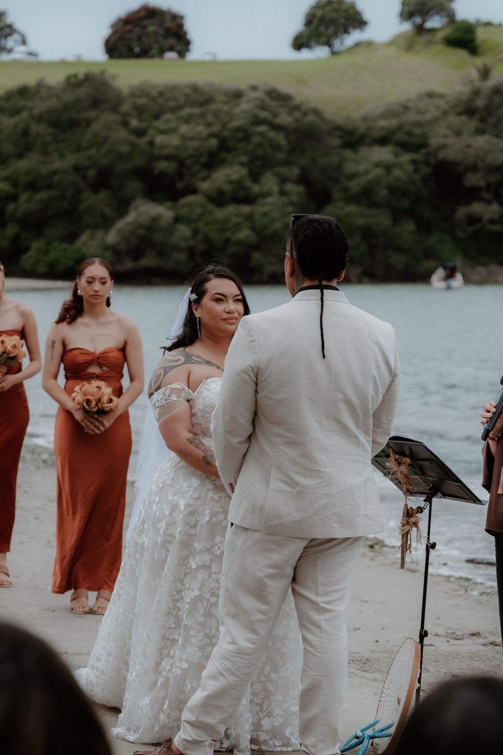 A beach wedding ceremony on the shoreline with bridesmaids in rust-colored dresses and a couple exchanging vows.