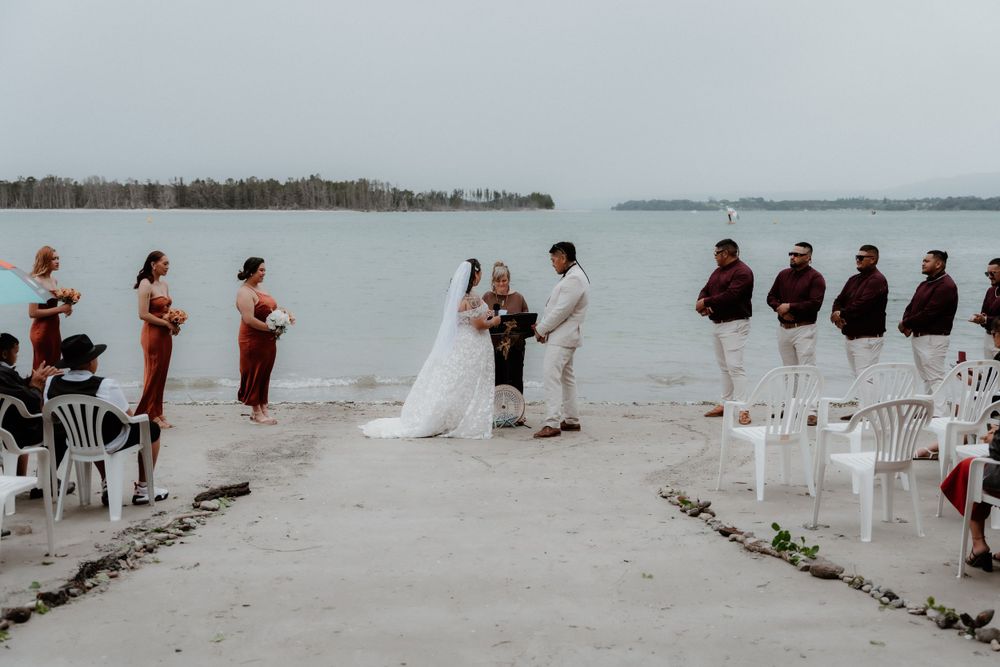 A beach wedding ceremony with guests seated in white chairs and the wedding party standing on either side.