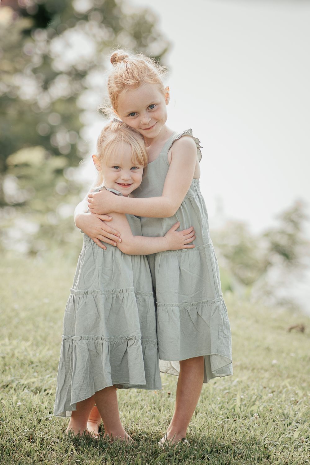 Two children in matching grey dresses share a sweet embrace in soft summer light.
