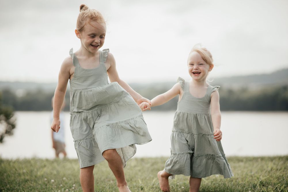 Two children in matching sage green dresses play and hold hands by a lake on a grassy hill.