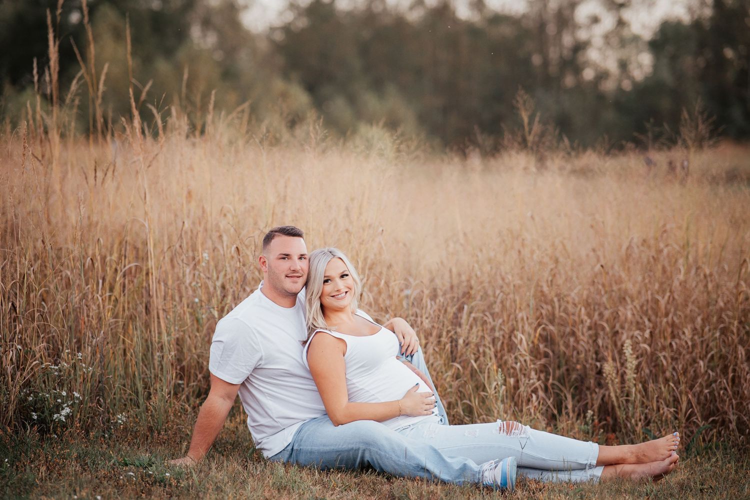 A couple sits together in a golden field of tall grass during sunset.