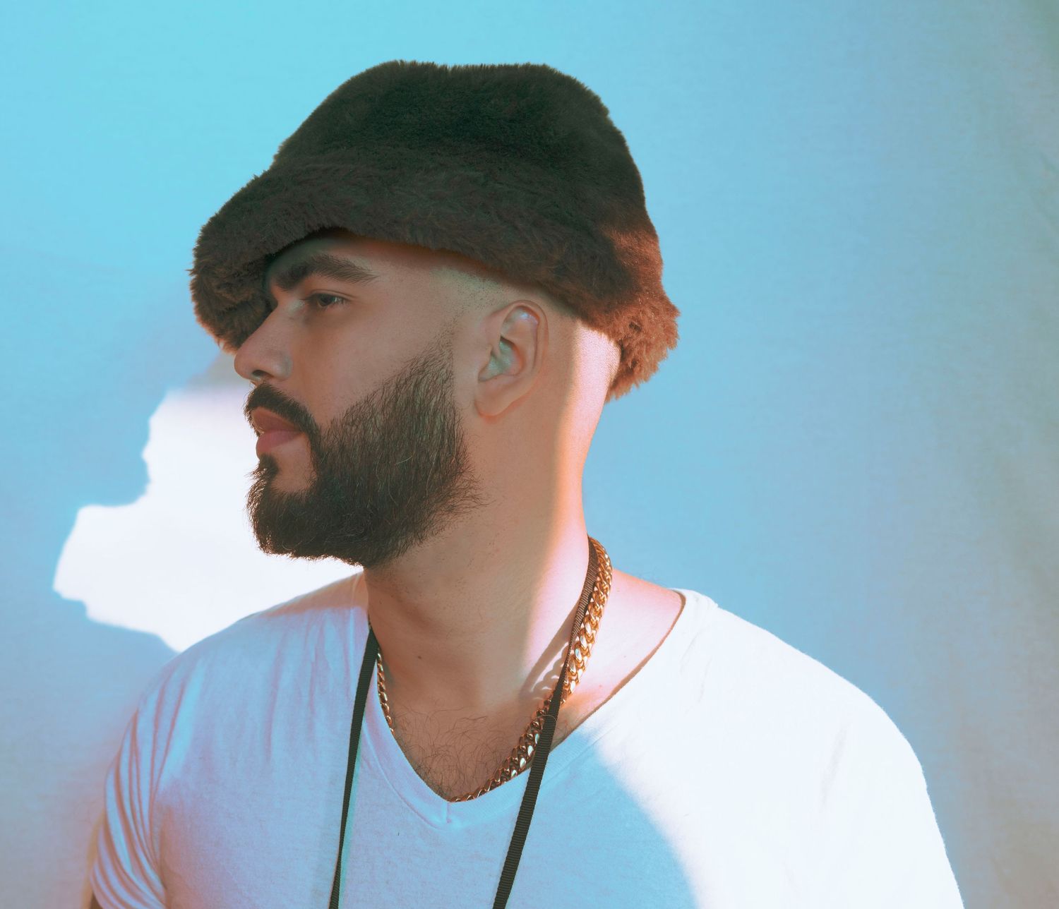 Stylish bearded man wearing a flat cap and white t-shirt with gold chain against a blue background.