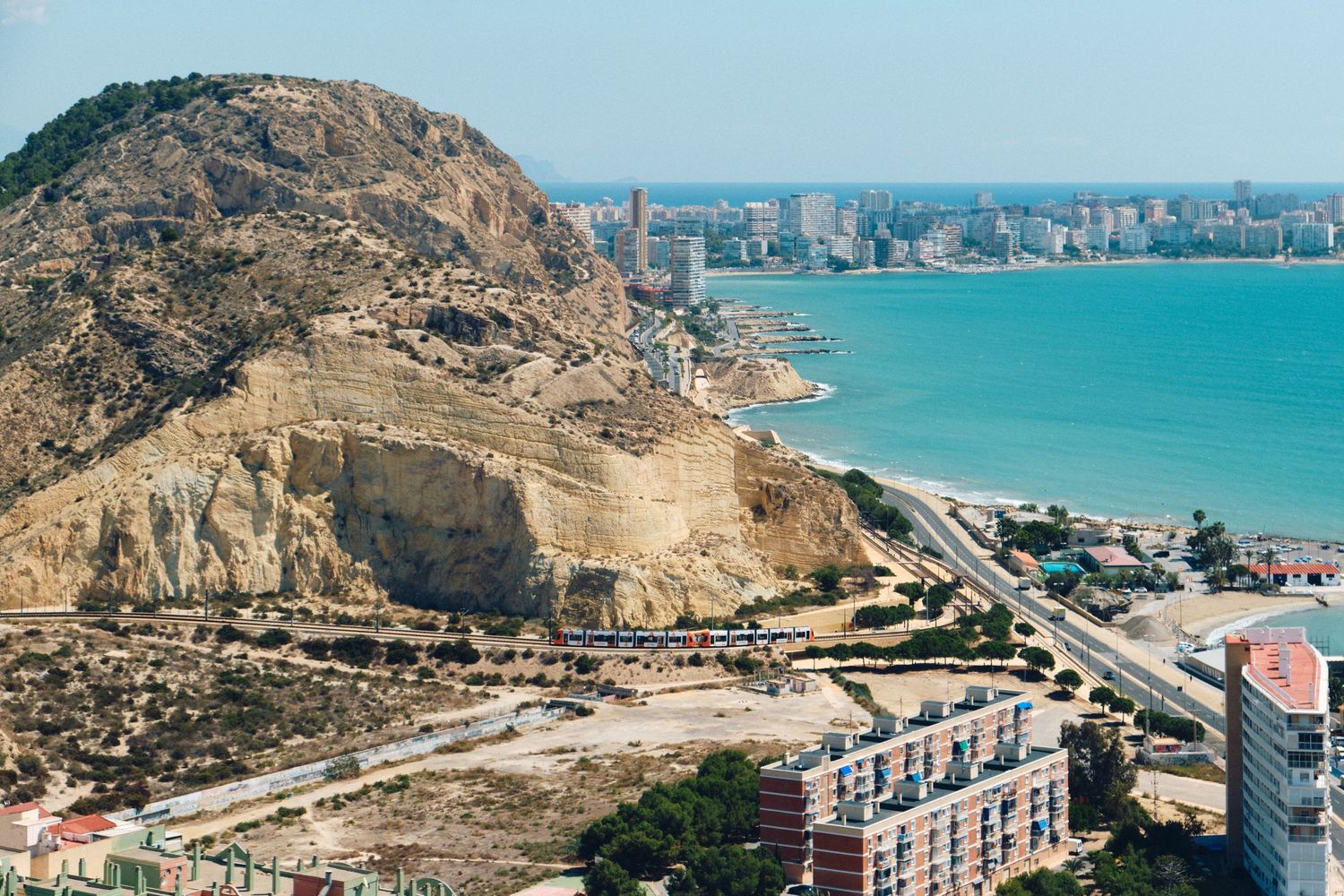 Aerial view of Alicante's coastline with mountains and buildings along the turquoise Mediterranean Sea.